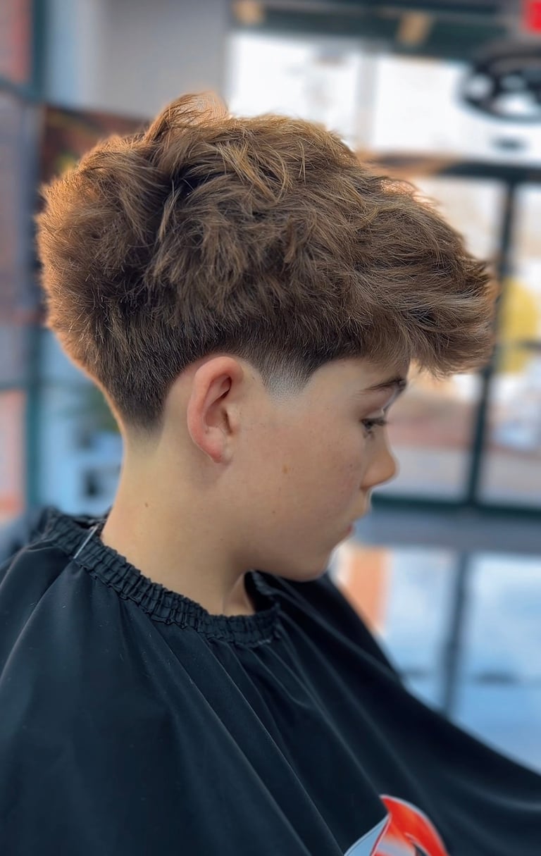 Young person with styled brown undercut hair with textured top, wearing black barber cape in salon setting