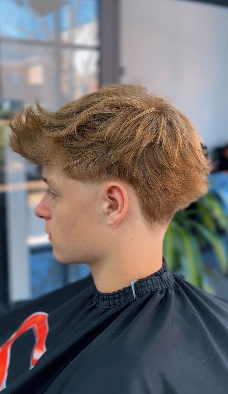 Side profile of a man with blonde textured hair styled with volume on top and faded sides, wearing a black cape in a salon setting