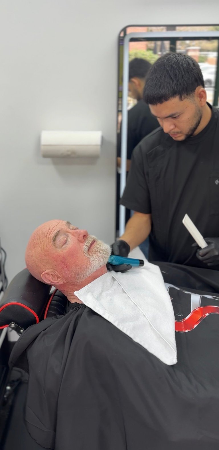 Young barber giving a haircut to an older man with a white beard in a barbershop setting