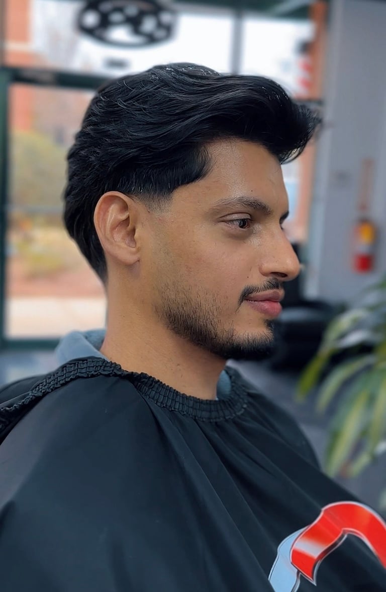 Man with styled black hair and facial hair wearing a black shirt, shown in profile view inside a barbershop setting.