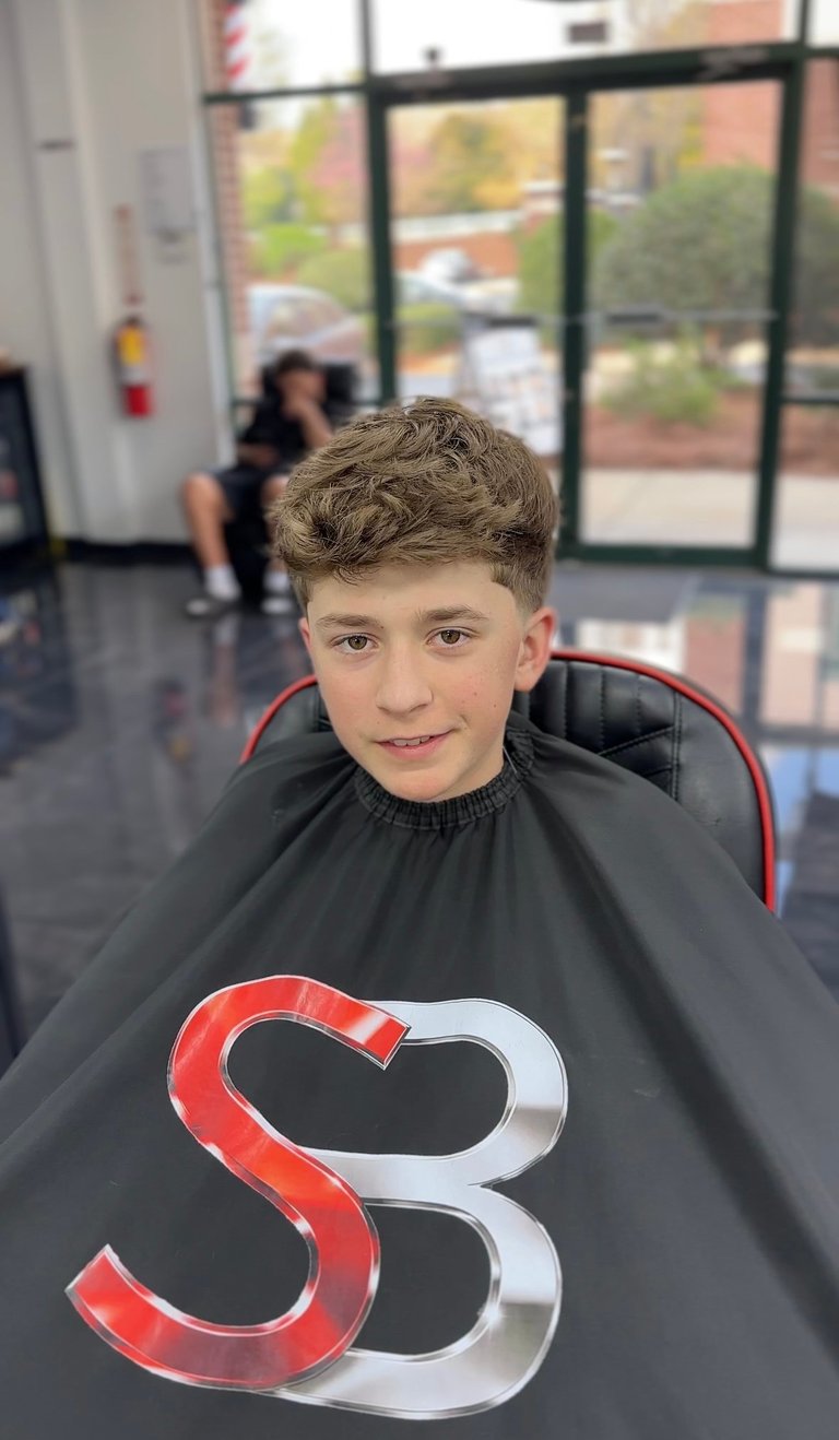 Young boy smiling at a barber shop wearing a black cape with a red and silver heart-shaped logo