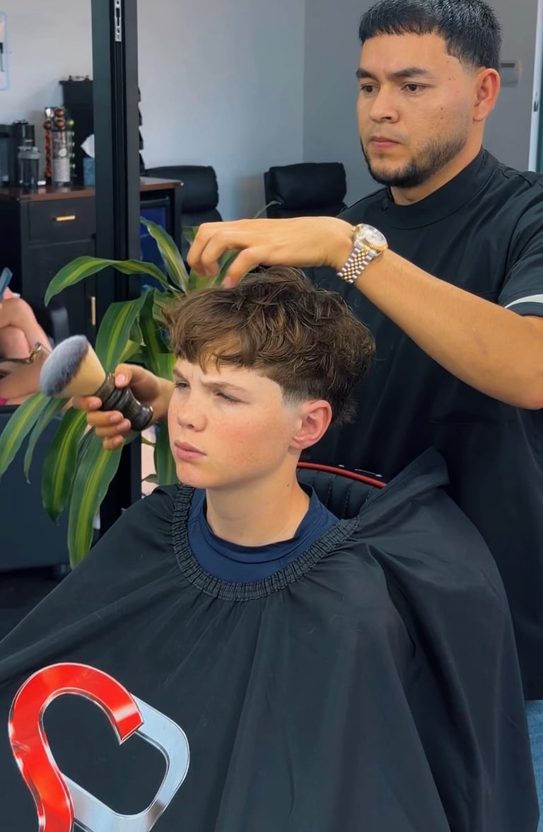 Barber styling a young boys hair with clippers in a salon. The boy wears a black cape while the barber uses styling tools on his brown hair.