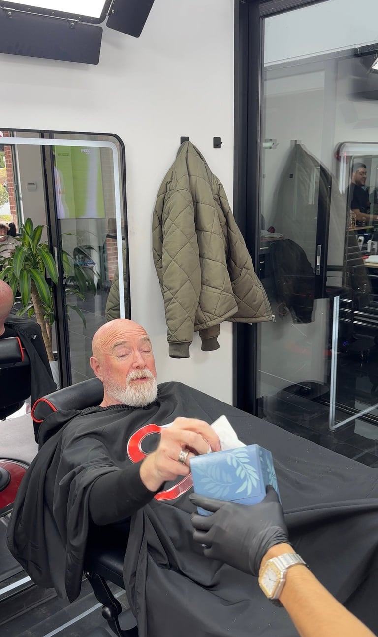 Older man with beard seated at table in salon, holding blue towel, with quilted jacket hanging on wall behind him